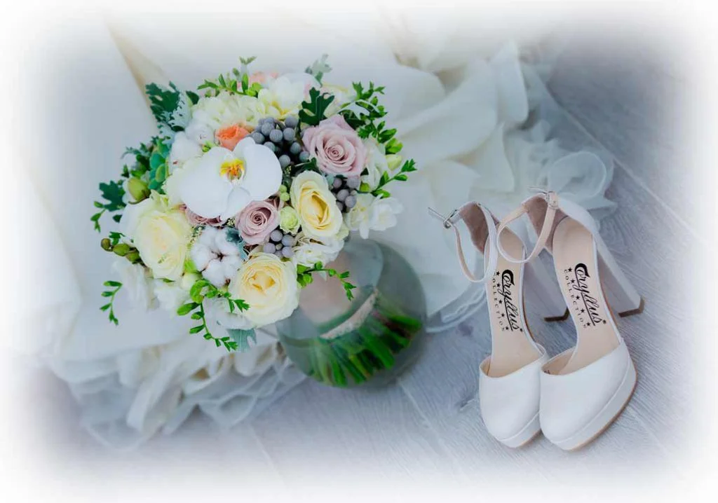 Ramo de novia de Nefertiti by Encina con rosas en tonos crema y rosa, orquídea blanca y detalles de follaje, acompañado de zapatos de novia blancos sobre un fondo elegante.